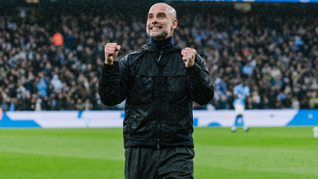 A coach in a black jacket celebrates with clenched fists on a football field with a cheering crowd in the background.