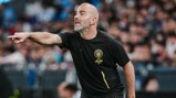 A coach with a shaved head, wearing a Manchester City training shirt, gestures assertively on the sideline during a match.