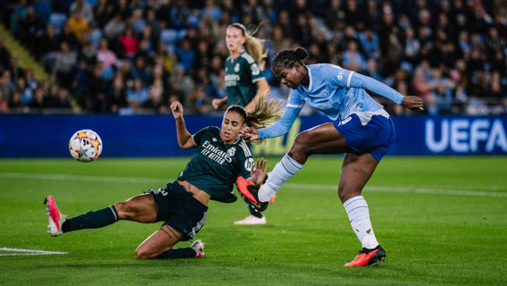 A soccer player in a Real Madrid jersey makes a tackle while a player in a Manchester City jersey takes a shot at the goal during a match.