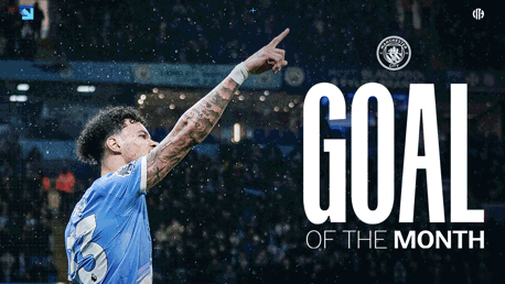 A football player wearing a Manchester City jersey points upwards in celebration of receiving the 'Goal of the Month' award. The Manchester City logo is visible in the background.