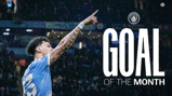 A football player wearing a Manchester City jersey points upwards in celebration of receiving the 'Goal of the Month' award. The Manchester City logo is visible in the background.