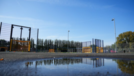Urban football field with metal fences and small goals, reflected in a puddle under a clear blue sky.