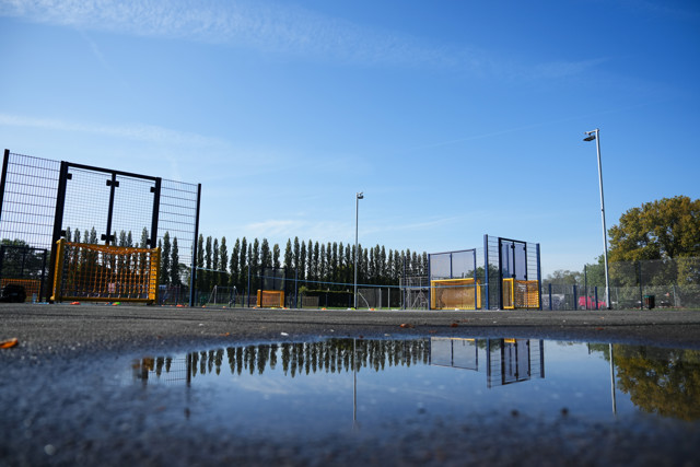 Urban football field with metal fences and small goals, reflected in a puddle under a clear blue sky.