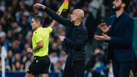 A football coach in a black jacket gestures towards an official holding a flag on the sideline during a match.