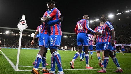 Crystal Palace players celebrating a goal during a football match against Fulham, evident from the corner flag displaying Fulham's logo. Players wear red and blue striped kits.