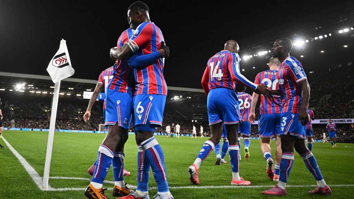 Crystal Palace players celebrating a goal during a football match against Fulham, evident from the corner flag displaying Fulham's logo. Players wear red and blue striped kits.