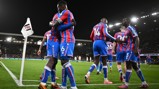 Crystal Palace players celebrating a goal during a football match against Fulham, evident from the corner flag displaying Fulham's logo. Players wear red and blue striped kits.