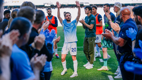 A football player wearing a light blue jersey is clapping in a guard of honor, surrounded by applauding players on a stadium field.