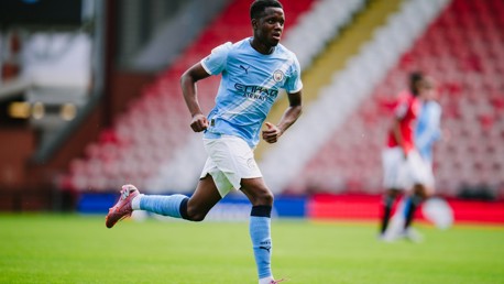 A Manchester City player is in action on the field during a football match. The player is wearing a light blue Manchester City kit and running across the green pitch.