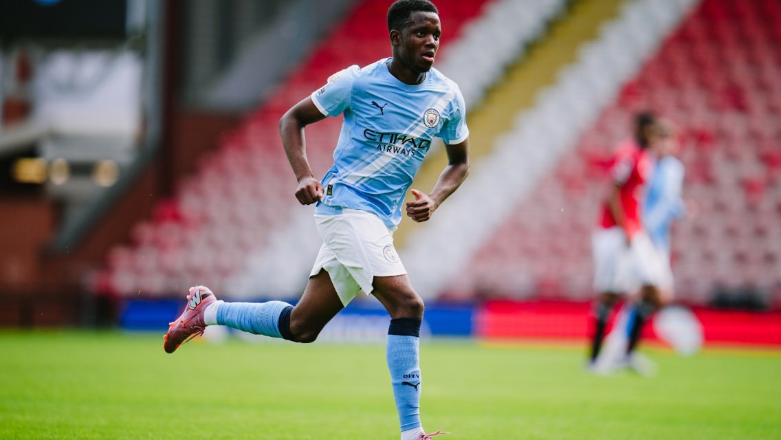A Manchester City player is in action on the field during a football match. The player is wearing a light blue Manchester City kit and running across the green pitch.