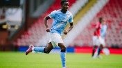 A Manchester City player is in action on the field during a football match. The player is wearing a light blue Manchester City kit and running across the green pitch.