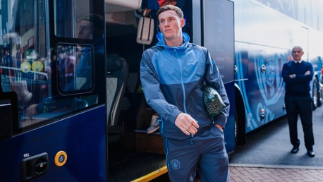 A person in a Manchester City tracksuit walks away from a team bus while holding a bag, with onlookers nearby.