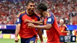 Two Spain football players celebrating during a match with one player pointing towards the other. The crowd is visible in the background. The City at the World Cup and FIFA logos are visible.