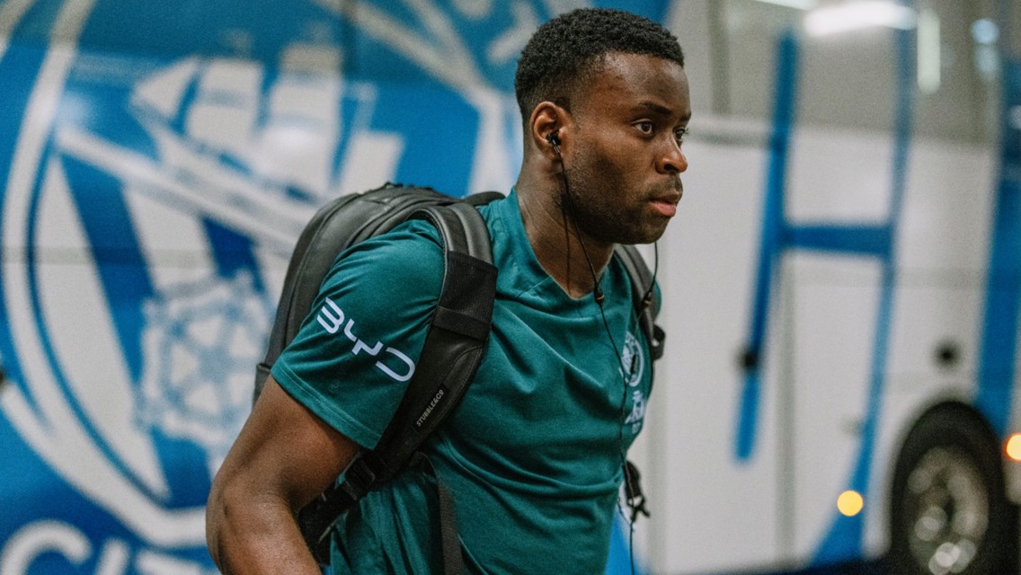 A person with a backpack and wearing a Manchester City shirt walks by the team bus.