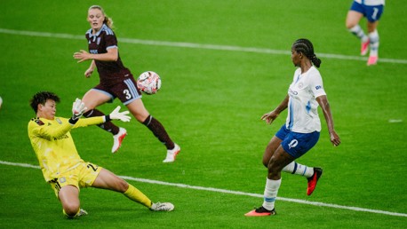 A soccer goalkeeper in a yellow kit is attempting to block a ball coming toward them during a match on a grassy field. Players in white and maroon jerseys are also visible.