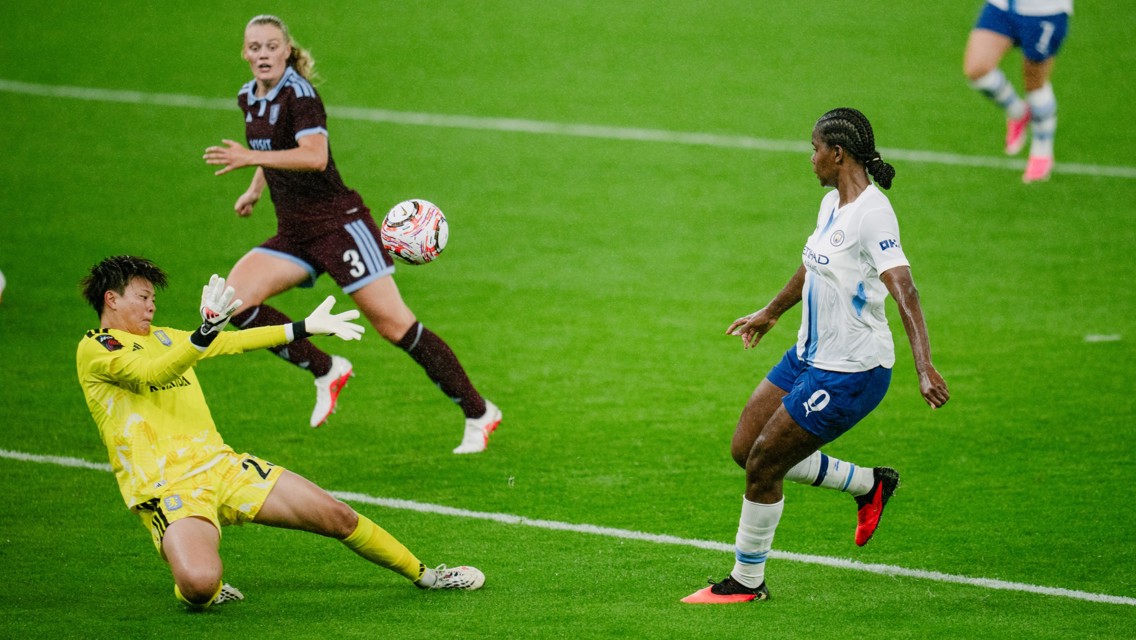 A soccer goalkeeper in a yellow kit is attempting to block a ball coming toward them during a match on a grassy field. Players in white and maroon jerseys are also visible.
