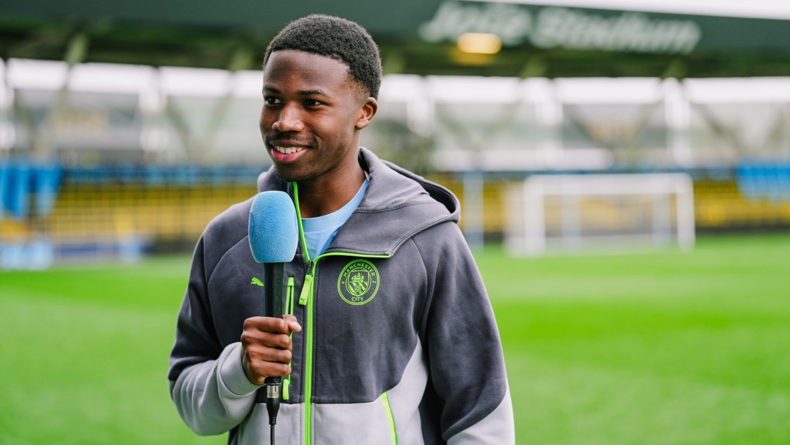 A person in a Manchester City jacket holds a microphone on a football field, standing in front of the goalposts.