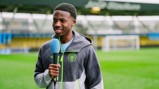 A person in a Manchester City jacket holds a microphone on a football field, standing in front of the goalposts.