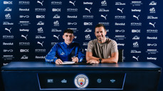 Two individuals at a signing table with Manchester City branding, including logos from sponsors like Etihad, Puma, and Revolut.