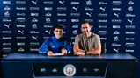 Two individuals at a signing table with Manchester City branding, including logos from sponsors like Etihad, Puma, and Revolut.