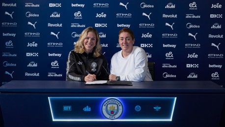 Two individuals at a Manchester City branded table during a contract signing event, with various sponsors in the background.
