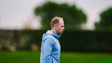 A person wearing a blue hoodie is outdoors with blurred face and green foliage in the background.