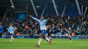 A Manchester City player in a light blue jersey celebrating a goal on the field with arms outstretched. Supporters in the background are cheering.