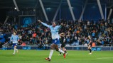 A Manchester City player in a light blue jersey celebrating a goal on the field with arms outstretched. Supporters in the background are cheering.