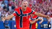 A football player in a red Norway jersey celebrating in a stadium. The jersey has the Norway flag design and the Manchester City logo appears in the corner.
