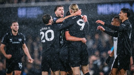 Manchester City players, including Bernardo Silva (#20) and Erling Haaland (#9), celebrate a goal in rainy weather. Faces are blurred.