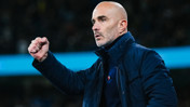 A bald man wearing a dark jacket and shirt makes a fist gesture, possibly encouraging or celebrating. The background appears to be a sports stadium.