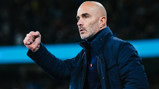 A bald man wearing a dark jacket and shirt makes a fist gesture, possibly encouraging or celebrating. The background appears to be a sports stadium.