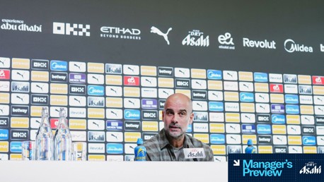 Press conference setting featuring branded backdrop with logos like Puma, Etihad. A person is speaking at the conference table with water bottles and a beverage can in front.