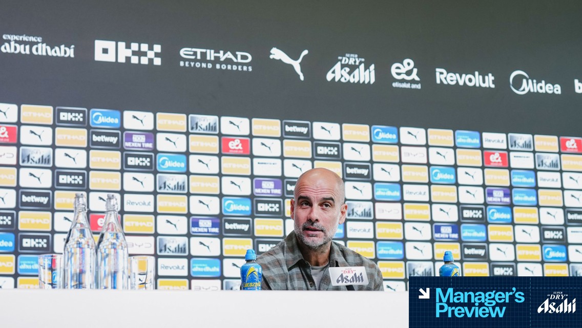 Press conference setting featuring branded backdrop with logos like Puma, Etihad. A person is speaking at the conference table with water bottles and a beverage can in front.