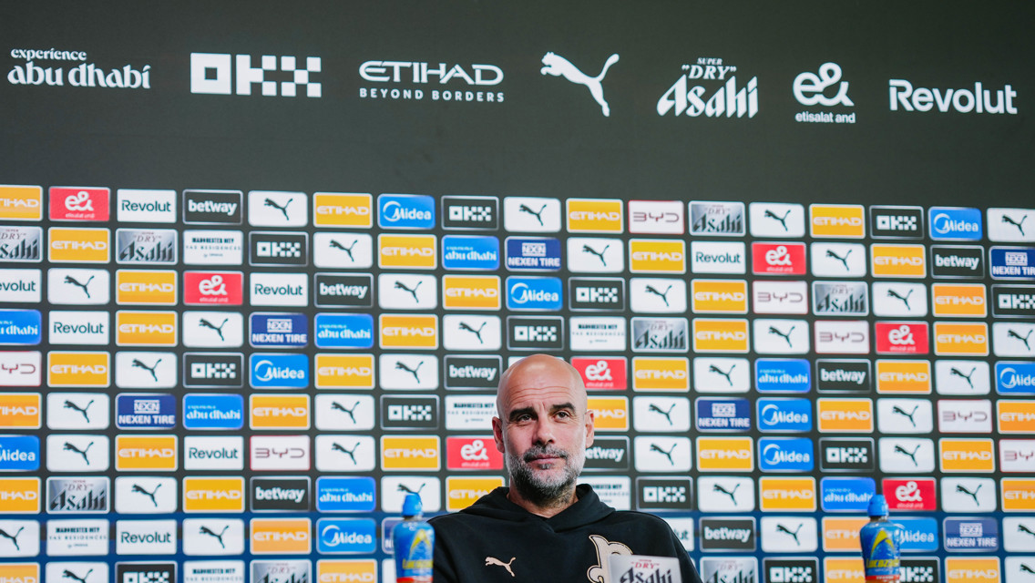 A press conference setting featuring a backdrop with multiple sponsor logos including Puma, Etihad, and Revolut. A partially blurred person is seated behind a desk with two bottles nearby.