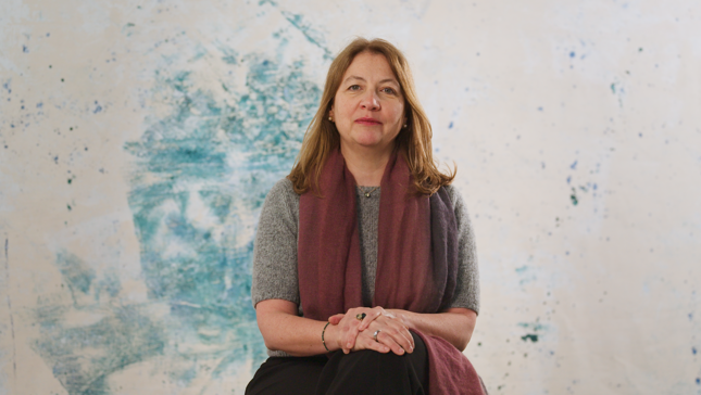 A woman with a blurred face sits cross-legged in front of a textured blue and white background, wearing a gray shirt and a burgundy scarf.