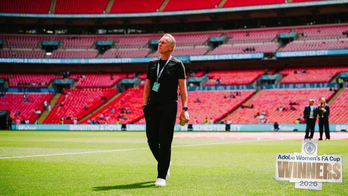 Person standing in Wembley stadium with empty seats. Brighton & Hove Albion and Manchester City banners in background. Adobe Women's FA Cup Winners 2026 logo visible.