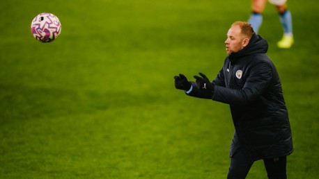 A football coach, possibly from Manchester City, wearing a Puma coat and gloves, is looking at a ball mid-air on a grassy field, with another player visible in the background.