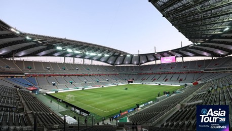 Wide angle view of a large, modern football stadium with empty seating and a green field. The venue is prepared for an event, possibly related to the Asia Tour 2026 as indicated by a logo in the corner.
