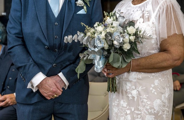 A close-up image of a wedding couple. The man is wearing a blue suit, white shirt, and light blue tie, while the woman is wearing a white lace dress. The woman is holding a bouquet of flowers.