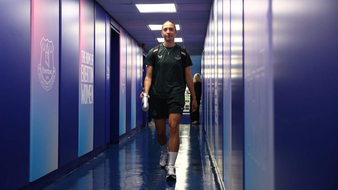 Person walking down a corridor in an Everton Women team facility, wearing sports attire and carrying a bottle. The walls have Everton logos and text.