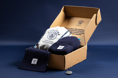 A membership pack featuring Manchester City fan items including a beanie, scarf, keychain, and a cloth bag with the Cityzens logo inside a cardboard box.