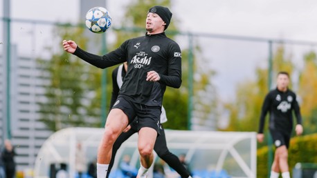 A soccer player in black training gear controls a ball during a practice session on a grass field, with teammates in the background.