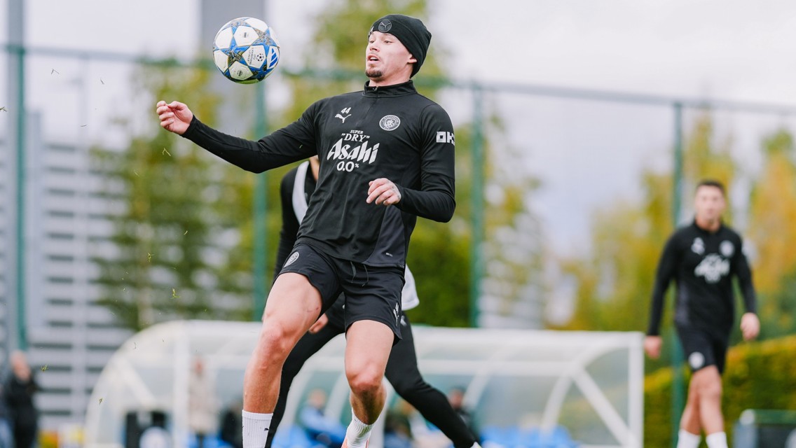 A soccer player in black training gear controls a ball during a practice session on a grass field, with teammates in the background.