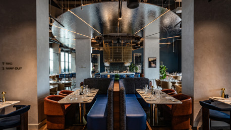 A modern restaurant interior featuring stylish booths, set tables with glassware and cutlery, and contemporary design elements such as hanging lights and reflective surfaces.