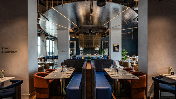 A modern restaurant interior featuring stylish booths, set tables with glassware and cutlery, and contemporary design elements such as hanging lights and reflective surfaces.