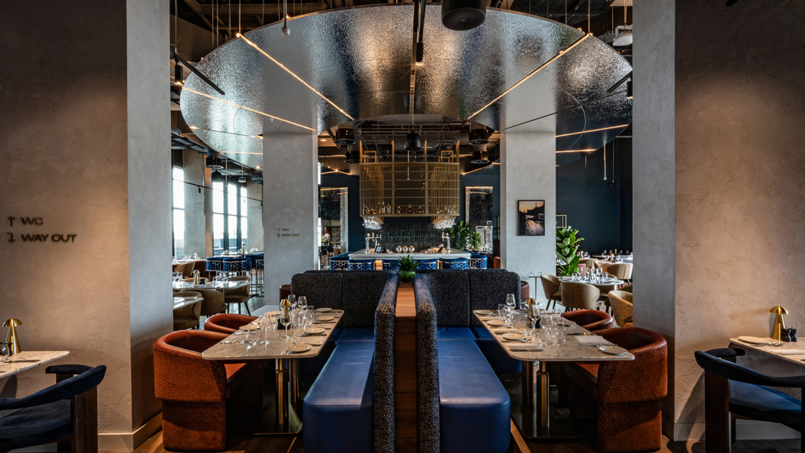 A modern restaurant interior featuring stylish booths, set tables with glassware and cutlery, and contemporary design elements such as hanging lights and reflective surfaces.