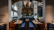 A modern restaurant interior featuring stylish booths, set tables with glassware and cutlery, and contemporary design elements such as hanging lights and reflective surfaces.
