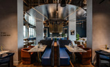 A modern restaurant interior featuring stylish booths, set tables with glassware and cutlery, and contemporary design elements such as hanging lights and reflective surfaces.