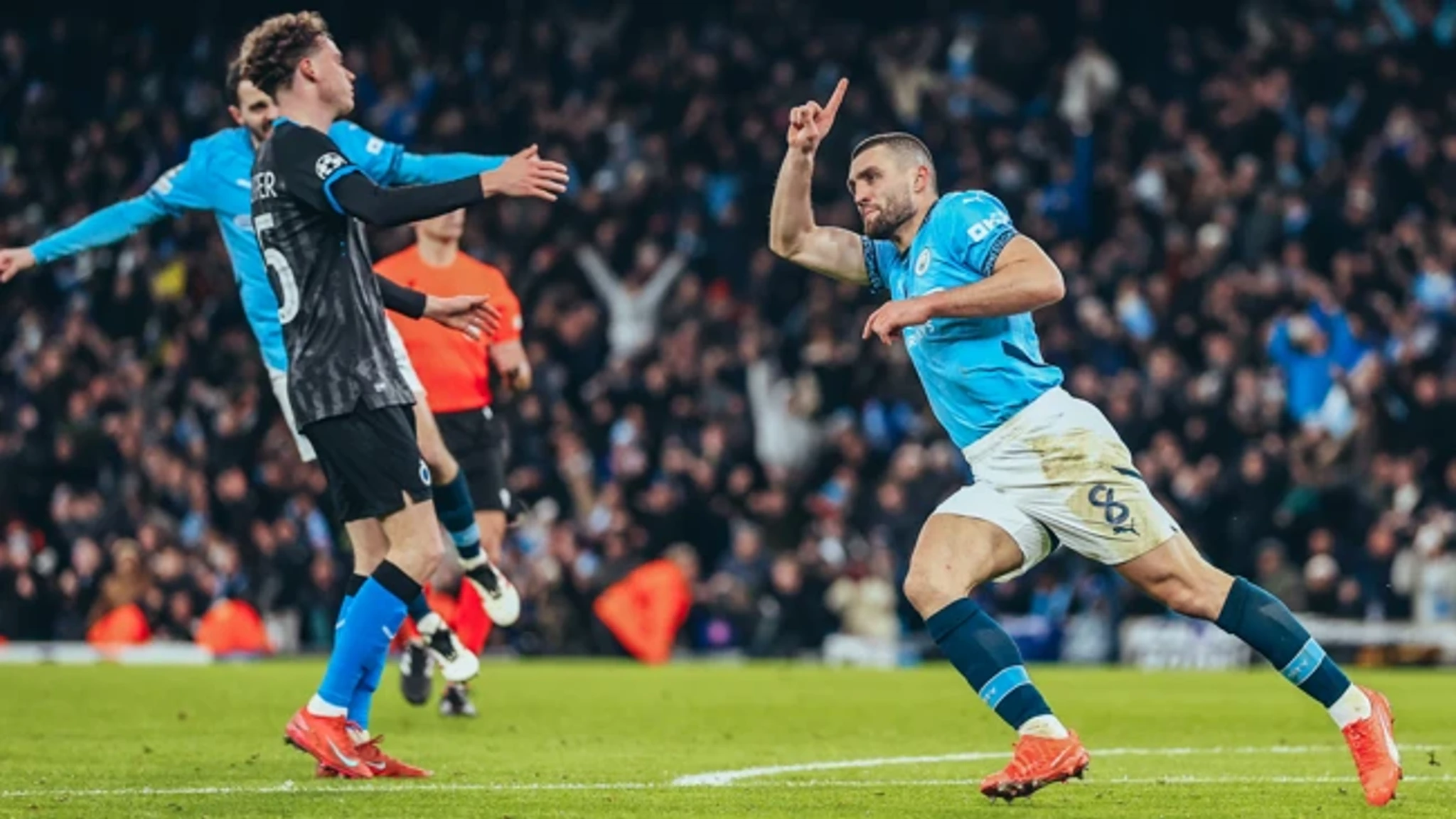 A football player in a blue Manchester City jersey is celebrating a goal with his right arm raised during a match. The referee and another player are visible in the background.